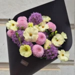 Hand holding a black-wrapped bouquet of pastel flowers including cream-yellow blooms, pink pom-poms, and purple alliums against a light stone background