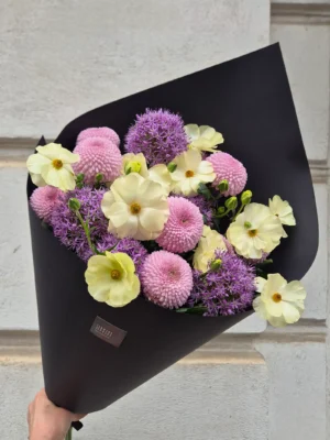 Hand holding a black-wrapped bouquet of pastel flowers including cream-yellow blooms, pink pom-poms, and purple alliums against a light stone background