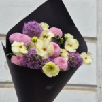 Bouquet of cream ranunculus, pink chrysanthemums, and purple alliums wrapped in black paper, held by a hand against a light wall.