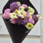 Bouquet of pale yellow ranunculus and cream flowers with purple allium and pink pompon mums in black wrapping, held by a hand against a gray wall.