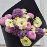 Bouquet of pale yellow flowers, purple alliums, and pink pompon mums wrapped in black paper, held against a light wall
