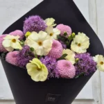 Bouquet of pale yellow daisies, pink chrysanthemums, and purple alliums wrapped in black paper cone.