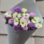 Bouquet of cream and pale yellow flowers with purple blooms, wrapped in gray paper and held by a hand against a stone wall background.