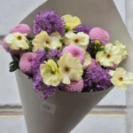 Bouquet of pale yellow ranunculus and cream petals with purple allium blooms, wrapped in gray paper and held by a hand