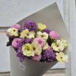 Bouquet of cream ranunculus, purple allium, and pink pompon chrysanthemums in gray wrapping, held by a hand against a light stone wall.