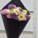 Bouquet of pale yellow and pink flowers wrapped in black paper, held by a hand against a light wall.