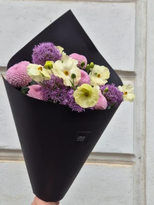Bouquet of pale yellow and pink flowers wrapped in black paper, held by a hand against a light wall.