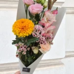 Bouquet of pink roses, yellow chrysanthemum, waxflowers and lisianthus held in light gray wrapping.