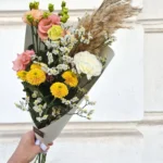 Bouquet of pastel pink roses, yellow chrysanthemums, white baby's breath, and tall pampas grass held in a white wrapping by a hand against a light stone wall