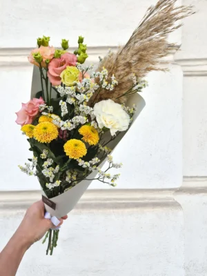 Bouquet of pastel pink roses, yellow chrysanthemums, white baby's breath, and tall pampas grass held in a white wrapping by a hand against a light stone wall