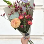 Hand-held bouquet with peach dahlias, pink ranunculus, purple lisianthus and statice wrapped in gray paper against a light wall.