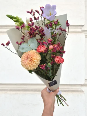 Hand-held bouquet with peach dahlias, pink ranunculus, purple lisianthus and statice wrapped in gray paper against a light wall.