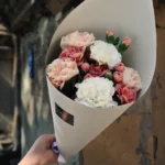 Bouquet of pink and white carnations wrapped in white paper, held by a hand outdoors against a stone wall.