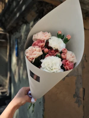 Bouquet of pink and white carnations wrapped in white paper, held by a hand outdoors against a stone wall.