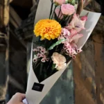 Hand holding a bouquet with pink roses, a bright yellow chrysanthemum, and small pink flowers in white wrapping against a textured wall.