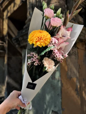 Hand holding a bouquet with pink roses, a bright yellow chrysanthemum, and small pink flowers in white wrapping against a textured wall.