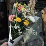 Bouquet of pink, yellow, and white flowers with dried grasses, wrapped in white paper, held against a stone wall. (outdoors)