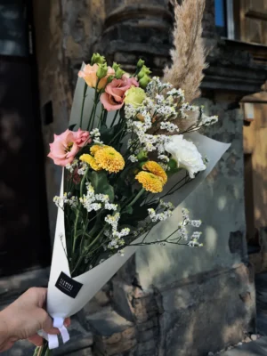 Bouquet of pink, yellow, and white flowers with dried grasses, wrapped in white paper, held against a stone wall. (outdoors)