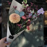 Bouquet of peach dahlia, pink ranunculus, purple statice, and white snapdragon wrapped in white paper, held against a stone wall backdrop.