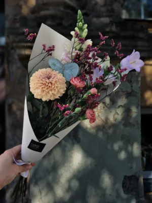Bouquet of peach dahlia, pink ranunculus, purple statice, and white snapdragon wrapped in white paper, held against a stone wall backdrop.