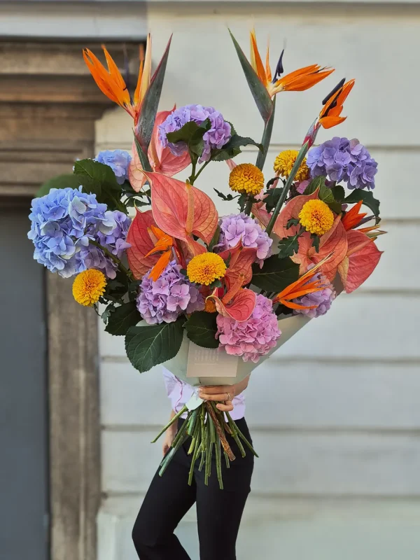 Person holding a large colorful bouquet featuring orange birds of paradise, purple hydrangeas, and yellow chrysanthemums in front of a neutral building.