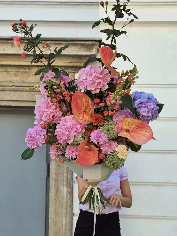 Person in a pastel shirt holding a large bouquet of pink hydrangeas, orange anthuriums, and purple dahlias in front of a building facade in a bridal-like arrangement.